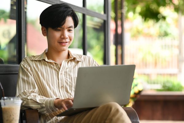 Smiling young Asian man sitting at outdoor cafe and using computer laptop.